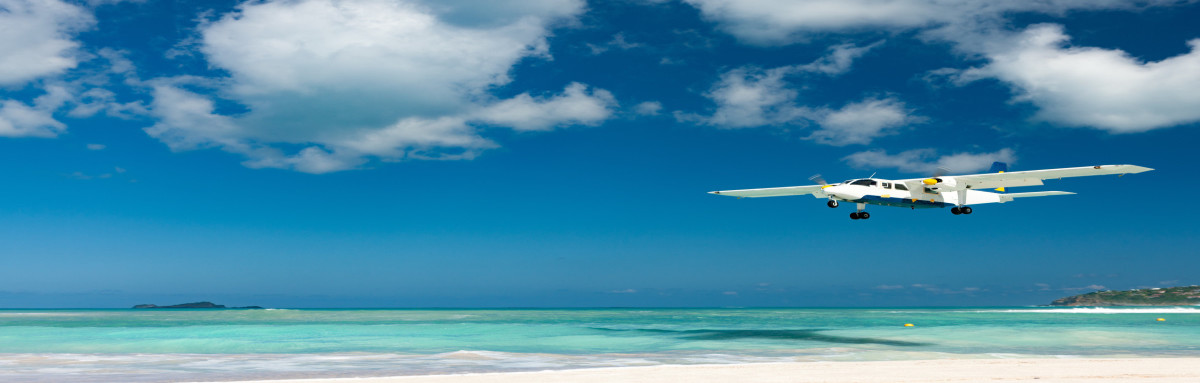 Small white plane flies over white sand beach and turquoise waters on a sunny day in St. Barts