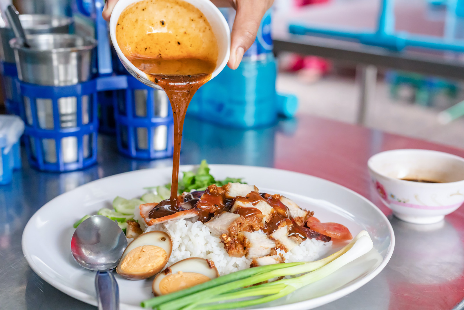 Red sauce being poured over chicken and rice with a hard boiled egg and a green spring onion on a plate with a metal spoon and cups with silverware in blue plastic holders and a white bowl with sauce on the side